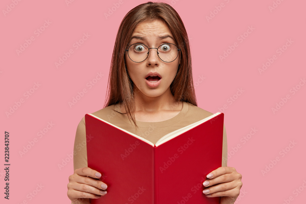 Stunned surprised teacher with book in hands, looks at pupil who made mess in classroom, wears transparent glasses, keeps jaw dropped, poses against pink background. Clever astonished student Stunned surprised teacher with book in hands, looks at pupil who made mess in classroom, wears transparent glasses, keeps jaw dropped, poses against pink background. Clever astonished student