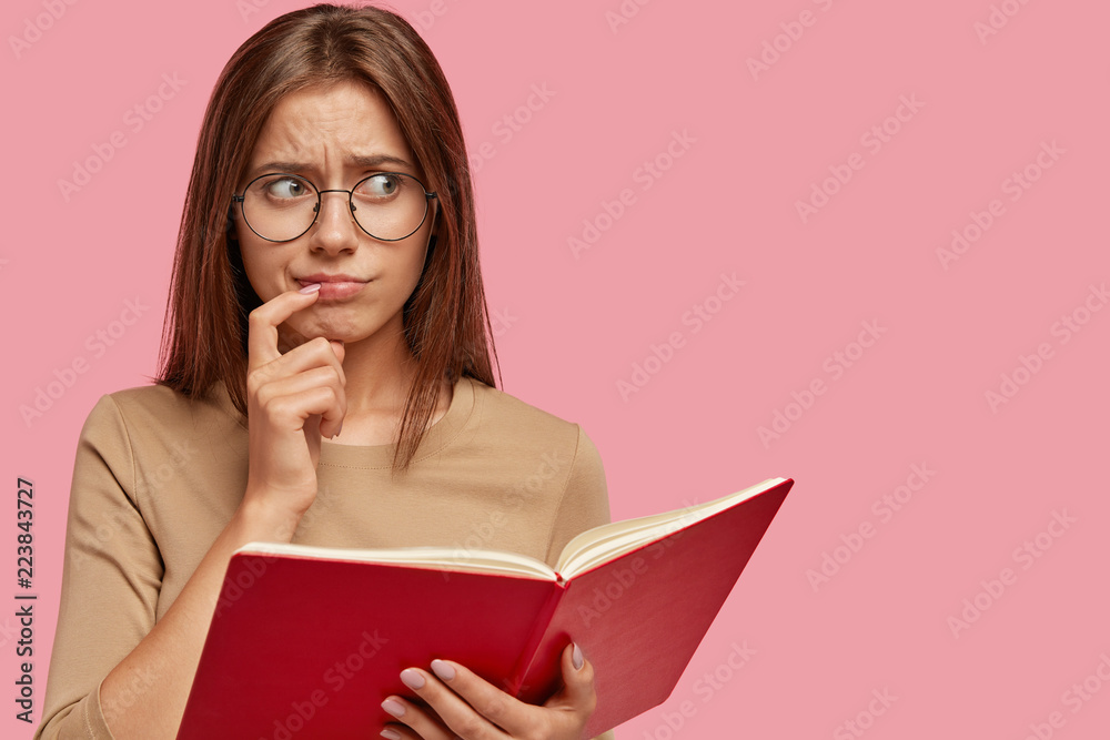 Frustrated puzzled schoolgirl keeps index finger near lips, carries red book, thinks in bewilderment how remember all material quickly, isolated over pink background with copy space for text