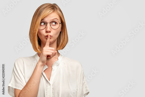 Shh, dont speak too loudly! Attractive appealing lady with short bobbed hairstyle, looks mysteriously aside, has secret expression, dressed in white casual clothes, isolated over studio background