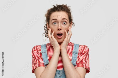 Bugged eyed terrifc girlfriend with curly hair, freckled skin, opens mouth widely, dressed in casual t shirt, denim overalls, stands against white background, being shocked with latest news.