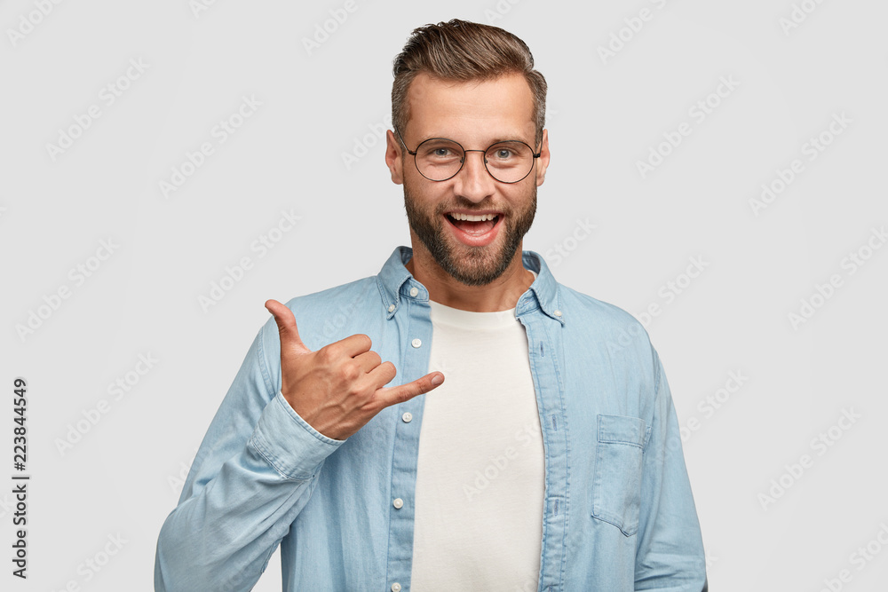 Friendly looking positive hipster with dark stubble, gestures indoor, shows shaka sign, being in high spirit, wears round spectacles for good vision, fashionable shirt, isolated on white wall