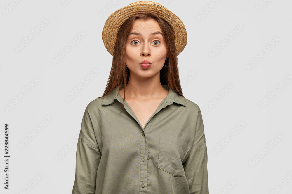 Beautiful youngster with pleasant appearance, keeps lips round, going to kiss someone, wears fashionable straw hat and shirt, poses against white background. Lovely girlfriend flirts with guy Beautiful youngster with pleasant appearance, keeps lips round, going to kiss someone, wears fashionable straw hat and shirt, poses against white background. Lovely girlfriend flirts with guy
