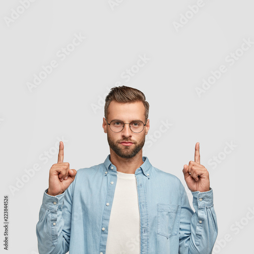 Photo of unshaven hipster dressed in casual shirt, points with both index fingers upwards. shows free space for your promotional content or advertisement. Lets go upstairs. Indoor vertical shot