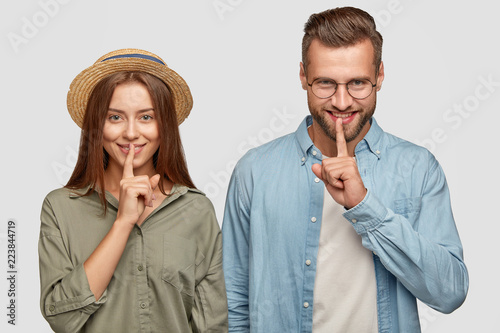 Shh, dont tell our secret. Positive young teenagers share rumours and gossip, keep index fingers on lips, ask to be quiet, isolated over white background. People, togetherness, secrecy concept photo