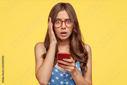 Photos Frightened stupefied young European girl keeps mouth opened, has fearful gaze at camera, recieves message reminding paying bills, stands against yellow background