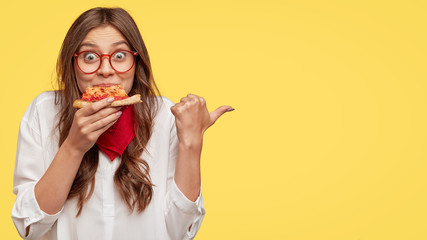 Lunch time concept. Attractive cheerful woman eats delicious Italian pizza, surprised with wonderful taste, indicates with thumb aside, shows where pizzeria situated, suggests to visit and have snack