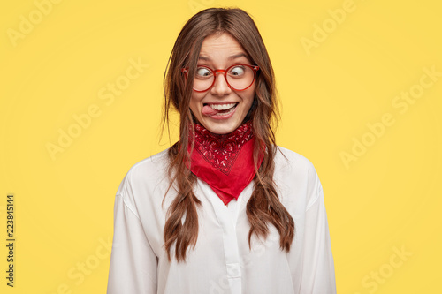 Fotografia Photo of crazy girlfriend makes funny face, crosses eyes and sticks out tongue, plays fool, doesnt want to be responsible, being in good mood, models indoor against yellow studio background