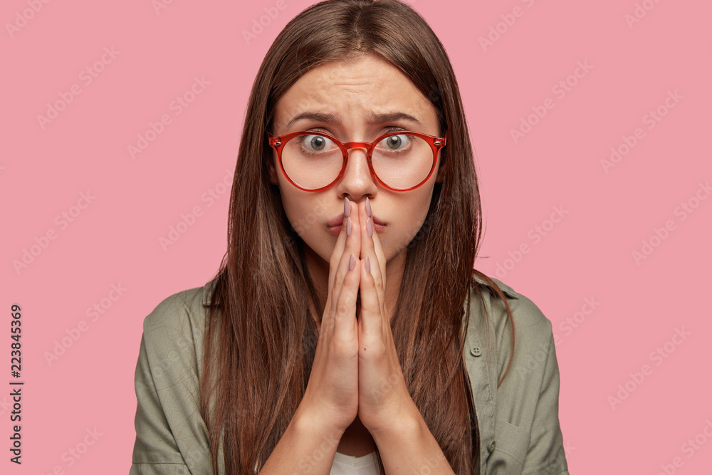 Photo of hopeful beautiful dark haired girl has miserable expression, keeps palms pressed together near lips, asks for forgiveness, poses against pink background. Cute woman beggs for something Photo of hopeful beautiful dark haired girl has miserable expression, keeps palms pressed together near lips, asks for forgiveness, poses against pink background. Cute woman beggs for something