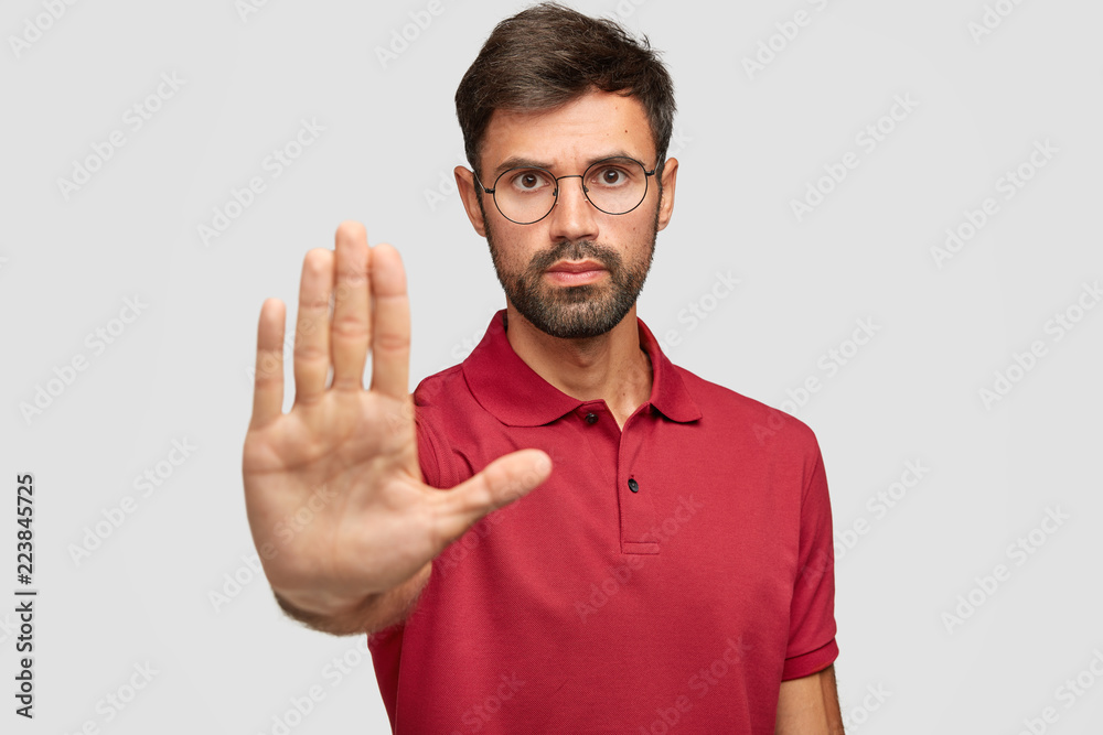 Fototapeta premium Concerned serious bearded man in round eyewear pulls palm towards camera, stops or warns you from making bad choice, shows enough or rejection gesture, isolated on white wall. Slow down, please!