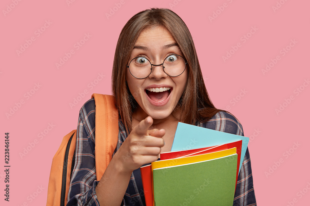 Close up shot of pupil with amazed expression, points directly into camera, carries rucksack, sees something amazing, isolated over pink background. People, studying, emotions and feeling concept Close up shot of pupil with amazed expression, points directly into camera, carries rucksack, sees something amazing, isolated over pink background. People, studying, emotions and feeling concept