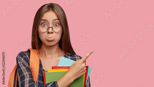 Photo of displeased offended Caucasian woman purses lower lip, carries rucksack, indicates with index finger at upper right corner, isolated over pink background. Youth and studying concept.