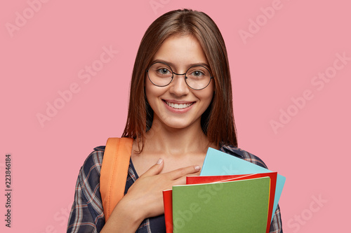 Thankful brunette woman with gentle smile, keeps hand on chest, expresses good feeling to friends and relatives, pleased to hear positive results of entrance exam carries bag and scientific literature