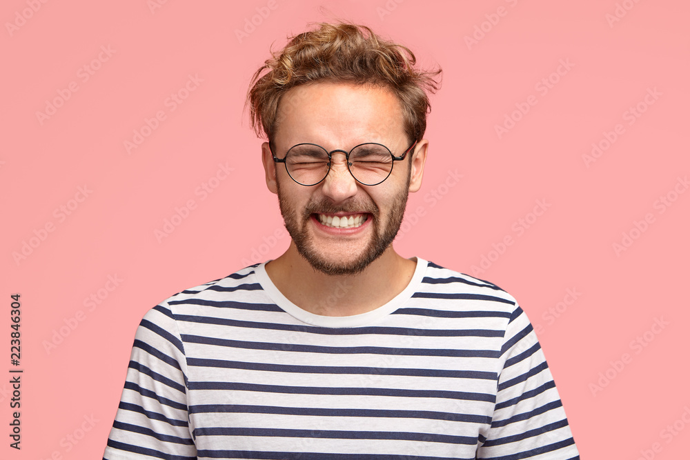Photo of attractive cheerful man laughs positively at camera, keeps eyes closed, dressed in casual striped t shirt, round spectacles, models in studio against pink background. Happy hipster indoor - obrazy, fototapety, plakaty 