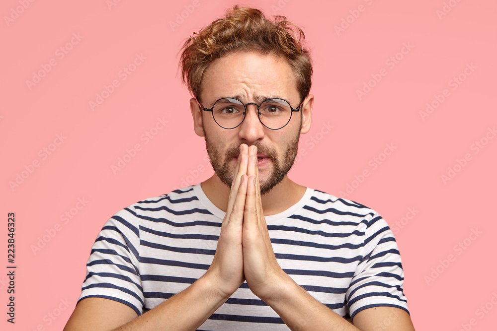 Miserable man with begging expression, asks for favor, keeps hands in praying gesture, wears stirped t shirt, stands against pink background. Please, cover me at work! Body language concept. Miserable man with begging expression, asks for favor, keeps hands in praying gesture, wears stirped t shirt, stands against pink background. Please, cover me at work! Body language concept.