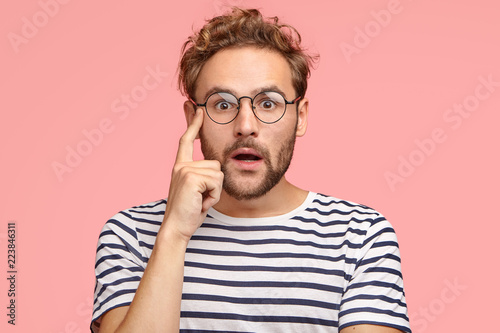 Portrait of astonished guy with appealing look looks with eyes full of disbelief, wears black and white striped casual t shirt, wonders as recieves unexpected proposal, isolated over pink studio wall photo