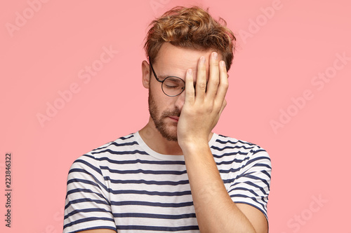 Tired overworked curly ordinary man keeps hand on face, closes eyes, feels sleepy after working all night, dressed in striped t shirt, isoated over pink studio wall. Fatigue freelancer needs rest.