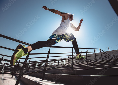 Fototapeta Naklejka Na Ścianę i Meble -  Handsome young muscular man in modern sport clothing run up the stairs outdoor at bright sunny day. Wide angle photo of a jogging man. Sport lifestyle