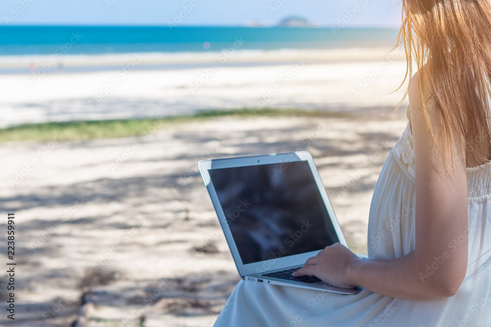 Young Asian woman using laptop in dress sitting on the beach,Girl Freelancer working on sea background