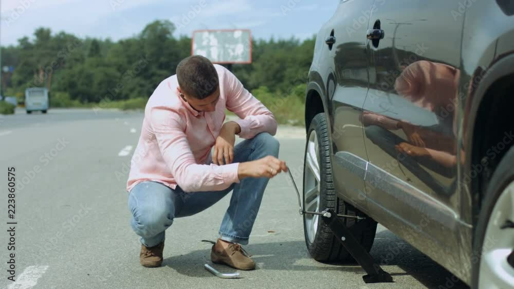 Handsome millennial male driver lifting his car on the jack to change flat tire on the road after tyre puncture during travel. Man with jack under his car lifting vehicle to replace tires on roadside.