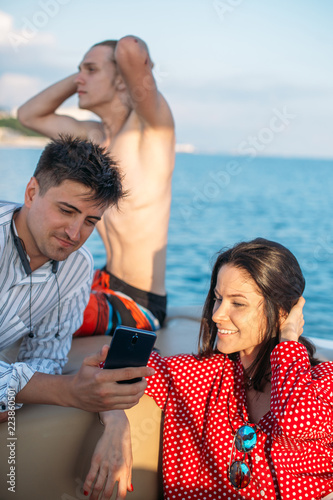 Two caucasian handsome men and one european woman having rest on sea boat during summer holyday marine trip. Man share with friends his photi on smartphone. photo