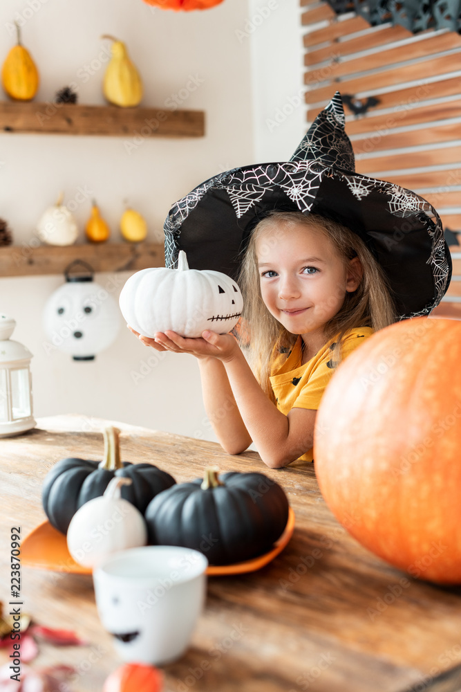 Fototapeta premium Cute little girl in witch costume sitting behind a table in Halloween theme decorated room, holding hand painted pumpkin and smiling. Halloween holiday concept.