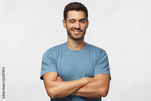 Handsome young man in blue t-shirt, with crossed arms smiling and winking, looking at camera isolated on gray background