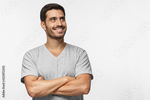 Portrait of young man with crossed arms and dreamy cheerful expression, thinking, isolated on gray background with copy space for your ads