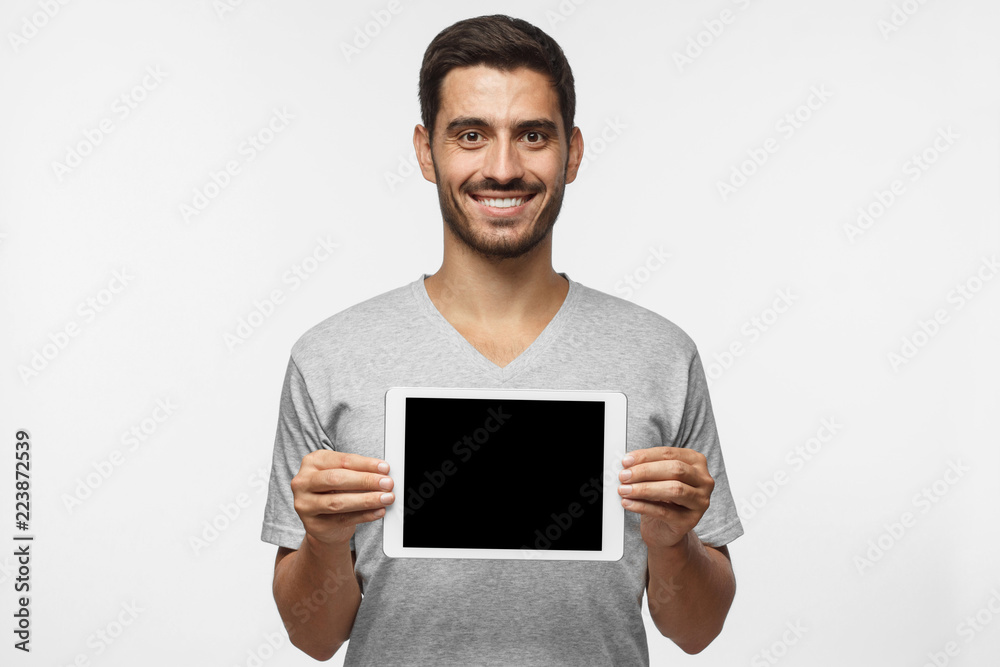 Young man isolated on gray background holding tablet and showing blank screen with happy smile as if advising product, service or app