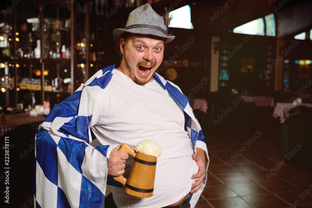 cheerful fat man with a mug of beer and with an oktoberfest flag in a  Bavarian hat on a pub background Stock Photo | Adobe Stock
