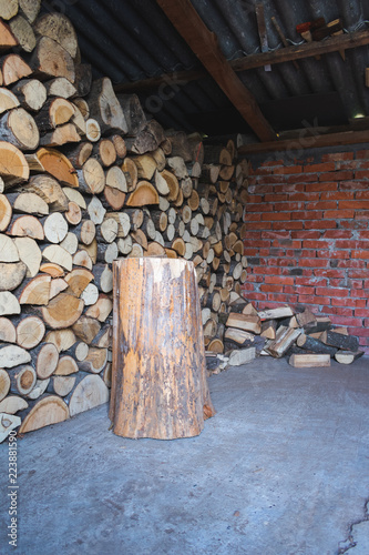 View of a stump for splitting logs and piles of logs or fire wood in a shed with a brick wall. Preparing for winter.