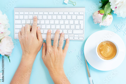 Flat lay home office workspace with someones hand typing white modern keyboard, notebook and peony flowers on blue background