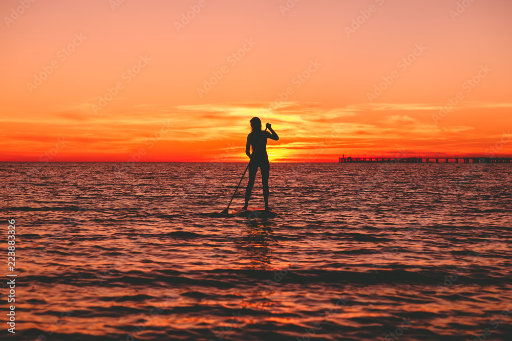 Silhouette of woman at stand up paddle board on a quiet sea with bright sunset or sunrise