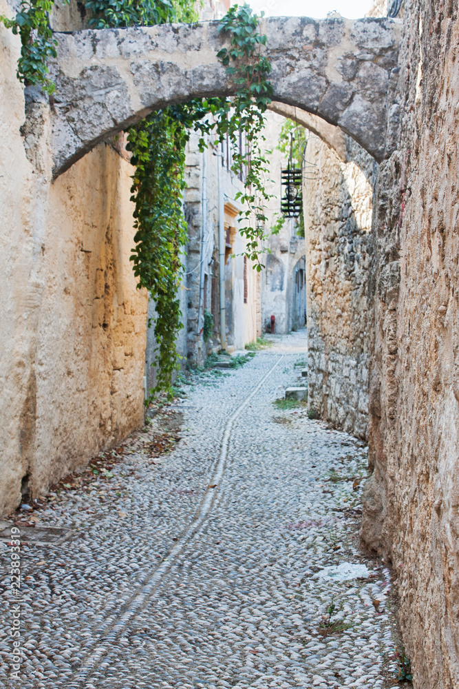 A medieval cobblestone alley in Rhodes Old Town (a UNESCO World Heritage Site), Greece, with arches and stone facades.