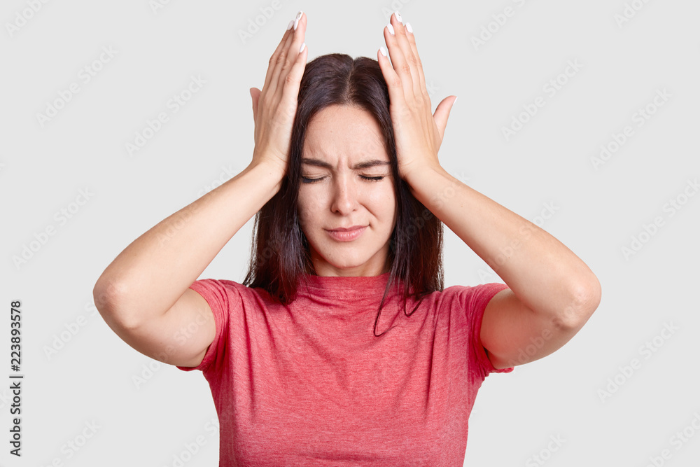 Photo of brunette woman with desperate look, keeps both hands on head, suffers from terrible headache, dressed in casual t shirt, poses against white background. People and tiredness conncept Photo of brunette woman with desperate look, keeps both hands on head, suffers from terrible headache, dressed in casual t shirt, poses against white background. People and tiredness conncept
