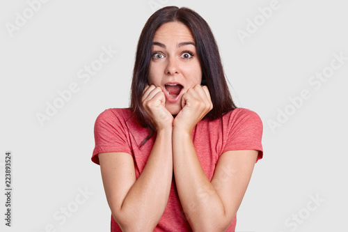 Slika na platnu Shocked beautiful brunette woman keeps mouth widely opened, holds chin with both hands, expresses great wonder, dressed in casual t shirt, poses against white background
