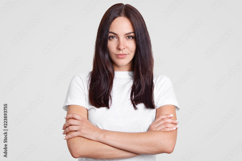 Charming self confident brunette woman with healthy skin, keeps arms folded, dressed in casual t shirt, listens information attentively, isolated over white background. People and seriousness Charming self confident brunette woman with healthy skin, keeps arms folded, dressed in casual t shirt, listens information attentively, isolated over white background. People and seriousness