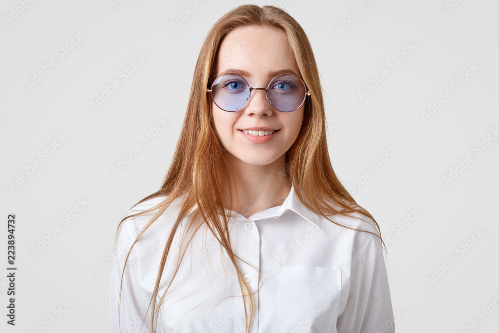Horizontal portrait of cheerful female student wears white shirt, trendy round shades, looks directly at camera, models in studio alone. People, lifestyle, positive emotions and fashion concept Horizontal portrait of cheerful female student wears white shirt, trendy round shades, looks directly at camera, models in studio alone. People, lifestyle, positive emotions and fashion concept