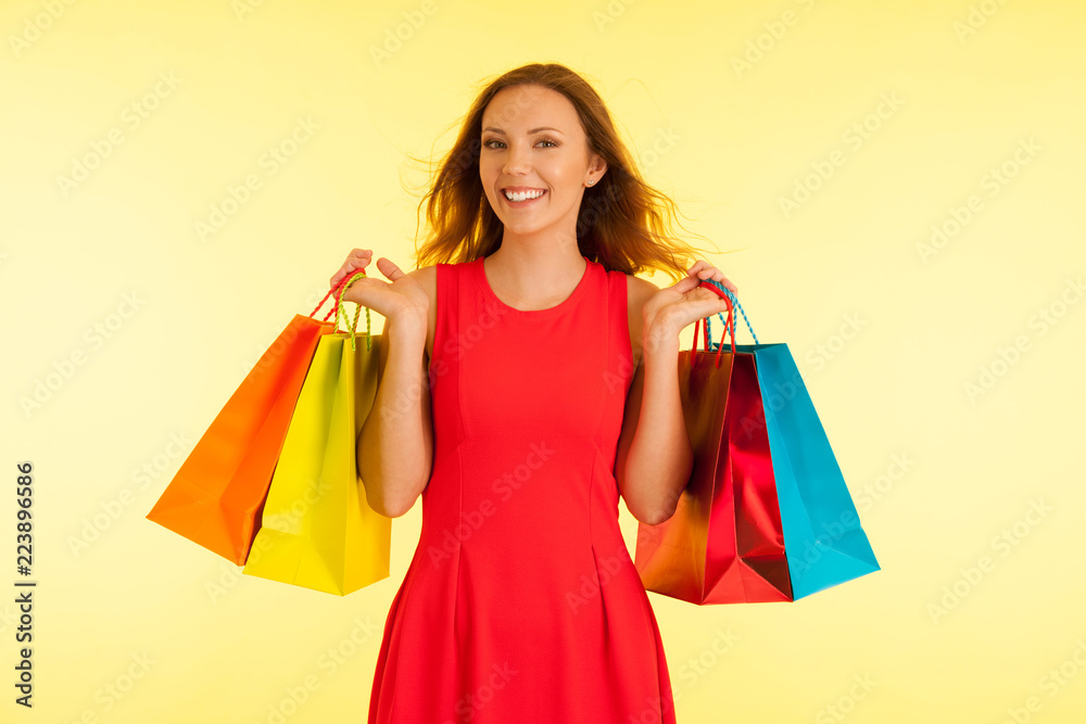 Beautiful young haapy woman in vibrant red dress holding shopping bags over yellow background