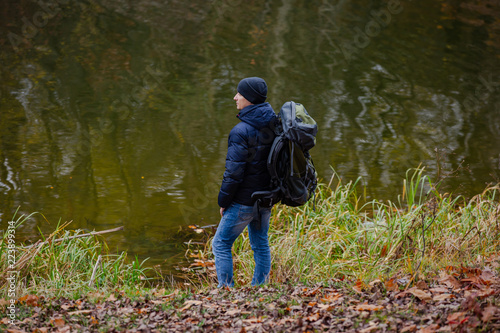 traveler stands with a backpack on his shoulders on the bank on the background of a lake in the autumn