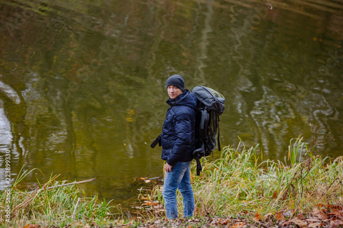 a tourist in a jacket and jeans looks at the camera near the river on the background of an autumn landscape © Vadim