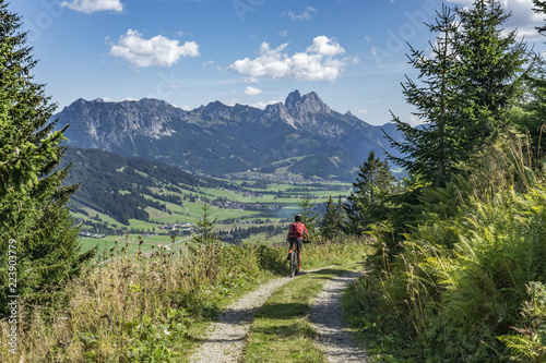 nice and active senior woman, riding her e-mountainbike in the Tannheim valley, Tirol, Austria with the village of Tannheim and famous summits Gimpel and Rote Flueh