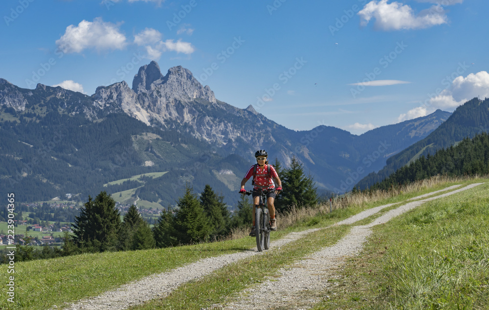 nice and active senior woman, riding her e-mountainbike in the Tannheim valley, Tirol, Austria with the village of Tannheim and famous summits Gimpel and Rote Flueh