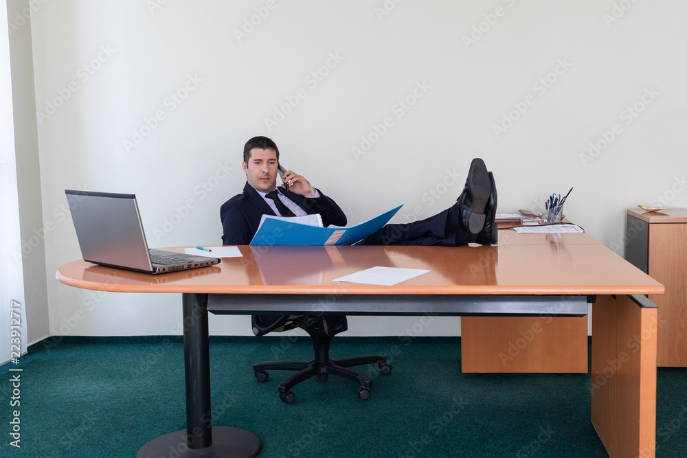 Portrait of young businessman sitting with his feet on the desk in the office talking on the mobile phone while verifying data from a file Portrait of young businessman sitting with his feet on the desk in the office talking on the mobile phone while verifying data from a file