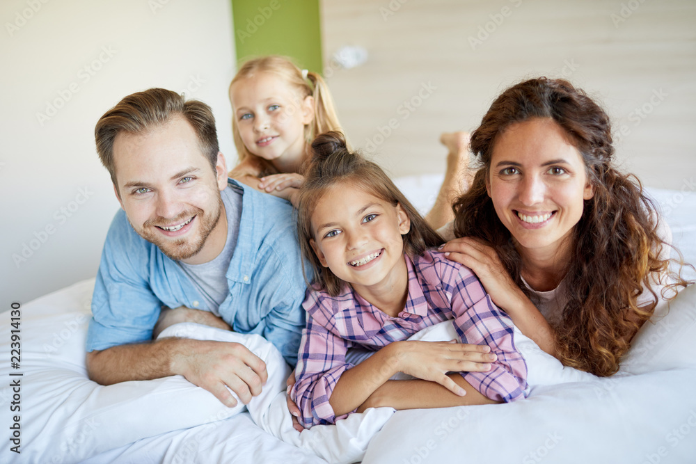 Cheerful family of mother, father and two cute little daughters lying on bed and looking at you