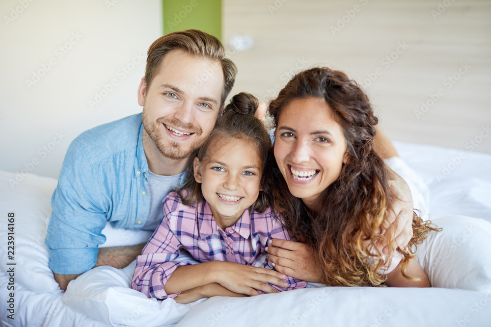 Young laughing restful couple and their daughter in casualwear lying on bed
