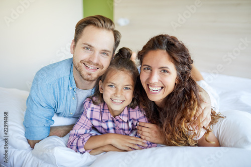 Young laughing restful couple and their daughter in casualwear lying on bed