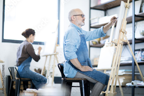 Mature man in workwear sitting in front of easel and sketching on paper sheet before painting
