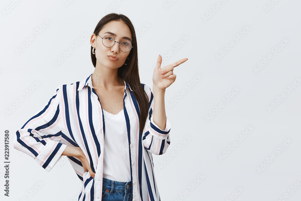 Horizontal shot of good-looking confident and happy female entrepreneur standing in self-assured feminine pose over gray background in stylish outfit, pointing right and folding lips cute Horizontal shot of good-looking confident and happy female entrepreneur standing in self-assured feminine pose over gray background in stylish outfit, pointing right and folding lips cute