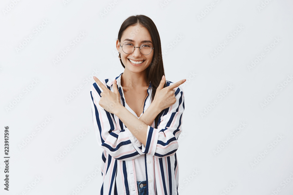 Woman satisfied with every chance. Charming happy female in glasses and striped blouse, crossing hands over chest and pointing left and right while smiling joyfully, posing over gray wall Woman satisfied with every chance. Charming happy female in glasses and striped blouse, crossing hands over chest and pointing left and right while smiling joyfully, posing over gray wall