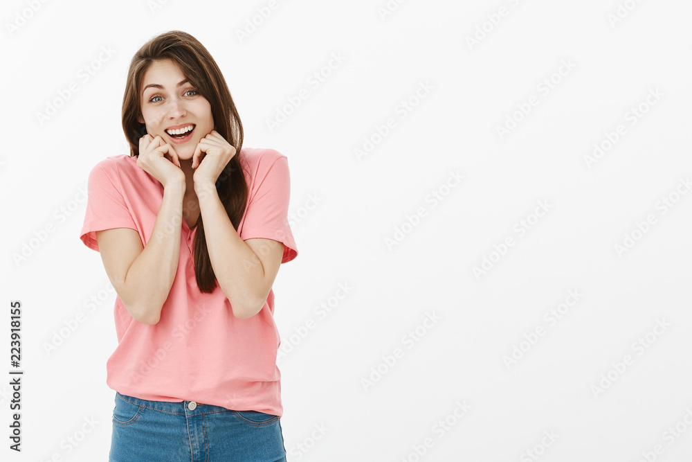 Indoor shot of excited attractive girlfriend with brown hair and beautiful smile holding hands near chin, being impressed and touched with amazing surprise, standing astonished over gray wall Indoor shot of excited attractive girlfriend with brown hair and beautiful smile holding hands near chin, being impressed and touched with amazing surprise, standing astonished over gray wall